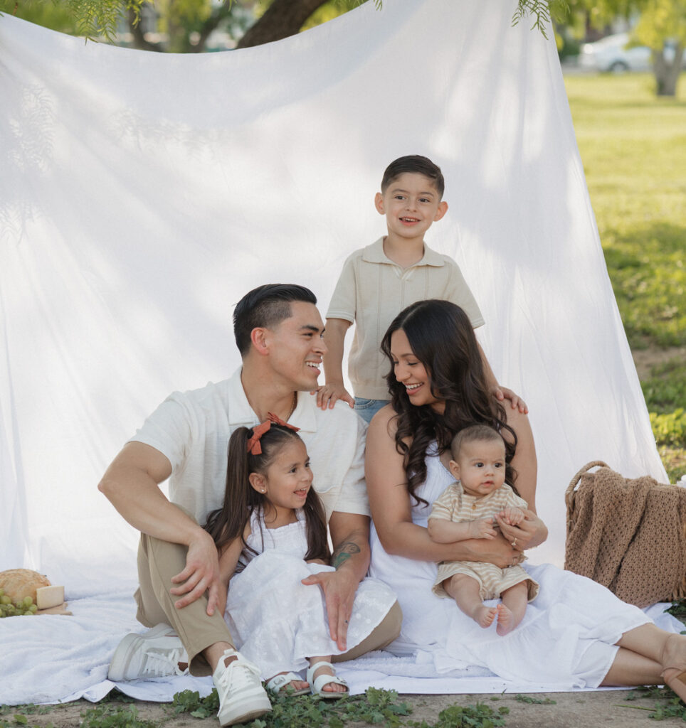 Cute family sits at a picnic at Mission Concepcion, with a white backdrop