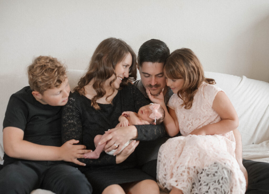 A cute family sits on the couch during an in home newborn session, everyone looking at the baby while mom looks at her next youngest's expression at the infant