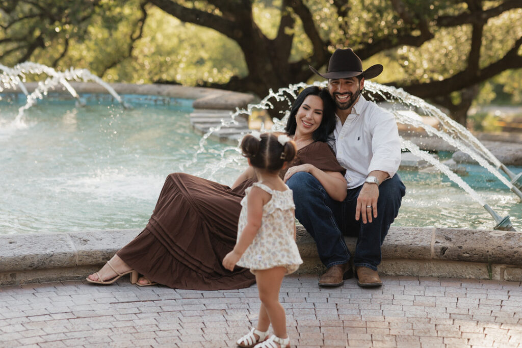 Young girl looks back at mom and dad, sitting in front of a fountain 