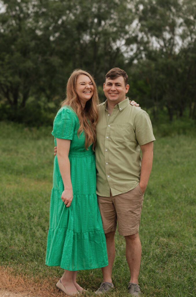 Husband and wife stand in a field, the backdrop of the trees and crescent bend nature park. She laughs as they stand, embracing each other, with one arm wrapped around each other.