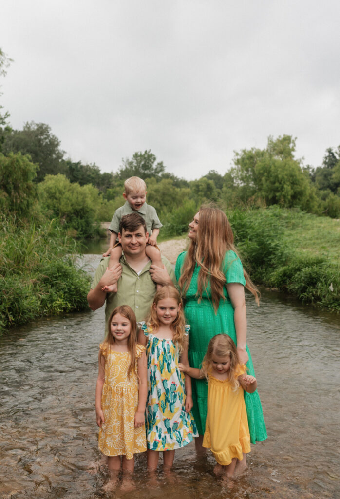 Husband, wife, and 4 kids, stand in the Cibolo Creek. The youngest, a boy, sits on his father's shoulders while his mother looks loving at him.
