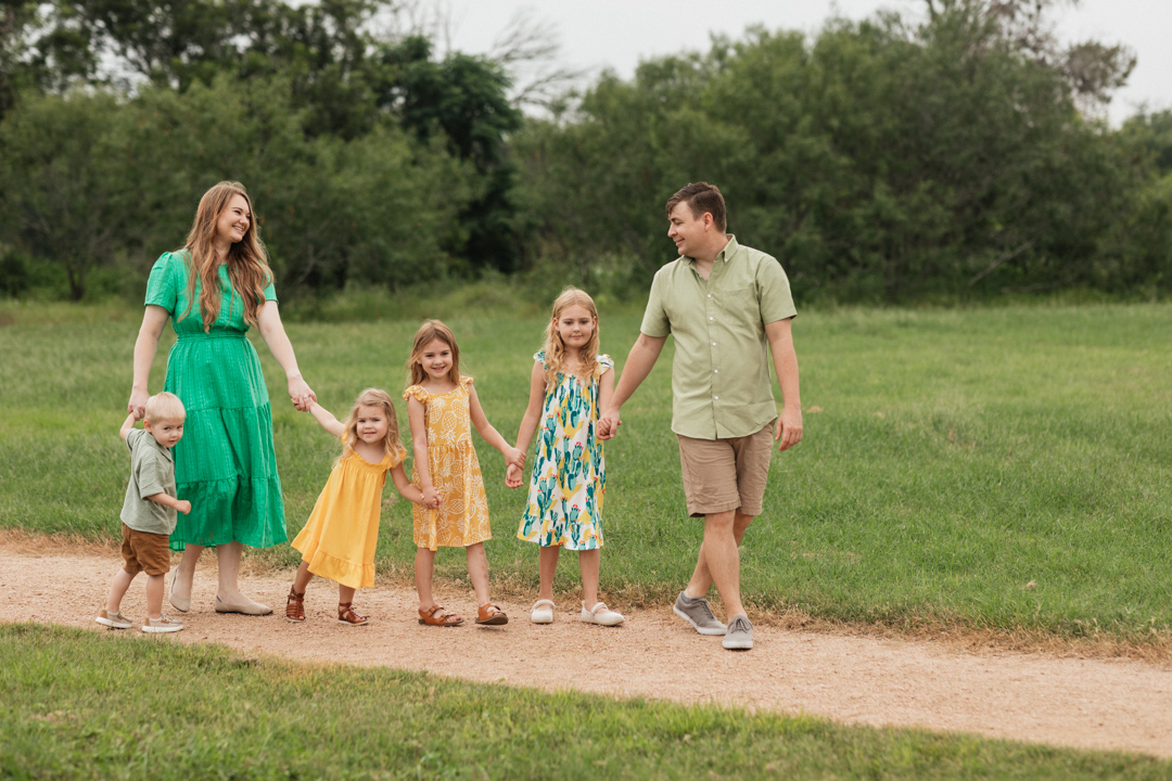 A family of four walks the trail through crescent bend nature park, holding hands in a chain with daddy leading the way.